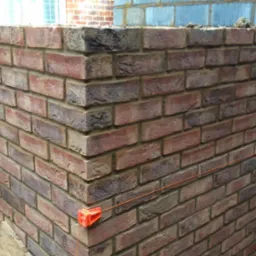 Wznoszenie narożnika ściany z czerwonej cegły, widoczne sznurki murarskie i zaprawa, w tle ściana z pustaków szarych.