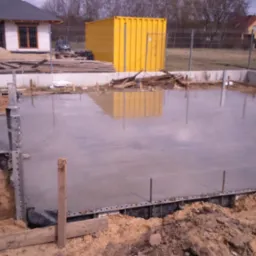 Świeżo wylany, gładki fundament pod budynek z widocznym żółtym kontenerem w tle, otoczony ziemią i drewnianymi elementami szalunku.