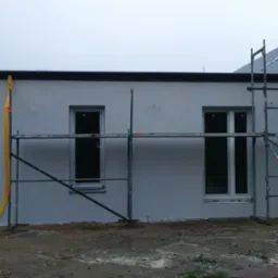 Świeżo otynkowany budynek w trakcie prac wykończeniowych, z widocznymi rusztowaniami i żółtymi rurami spustowymi do gruzu.