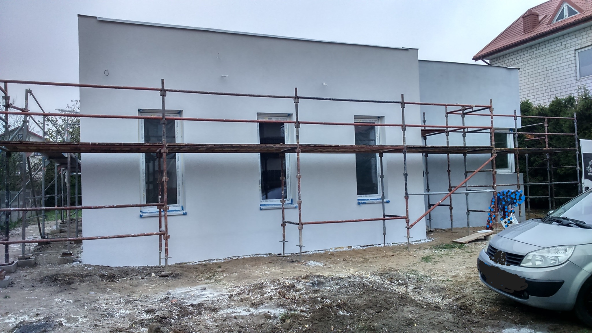 Świeżo otynkowana ściana domu jednorodzinnego w jasnym odcieniu szarości, z widocznymi rusztowaniami i oknami zabezpieczonymi taśmą malarską.