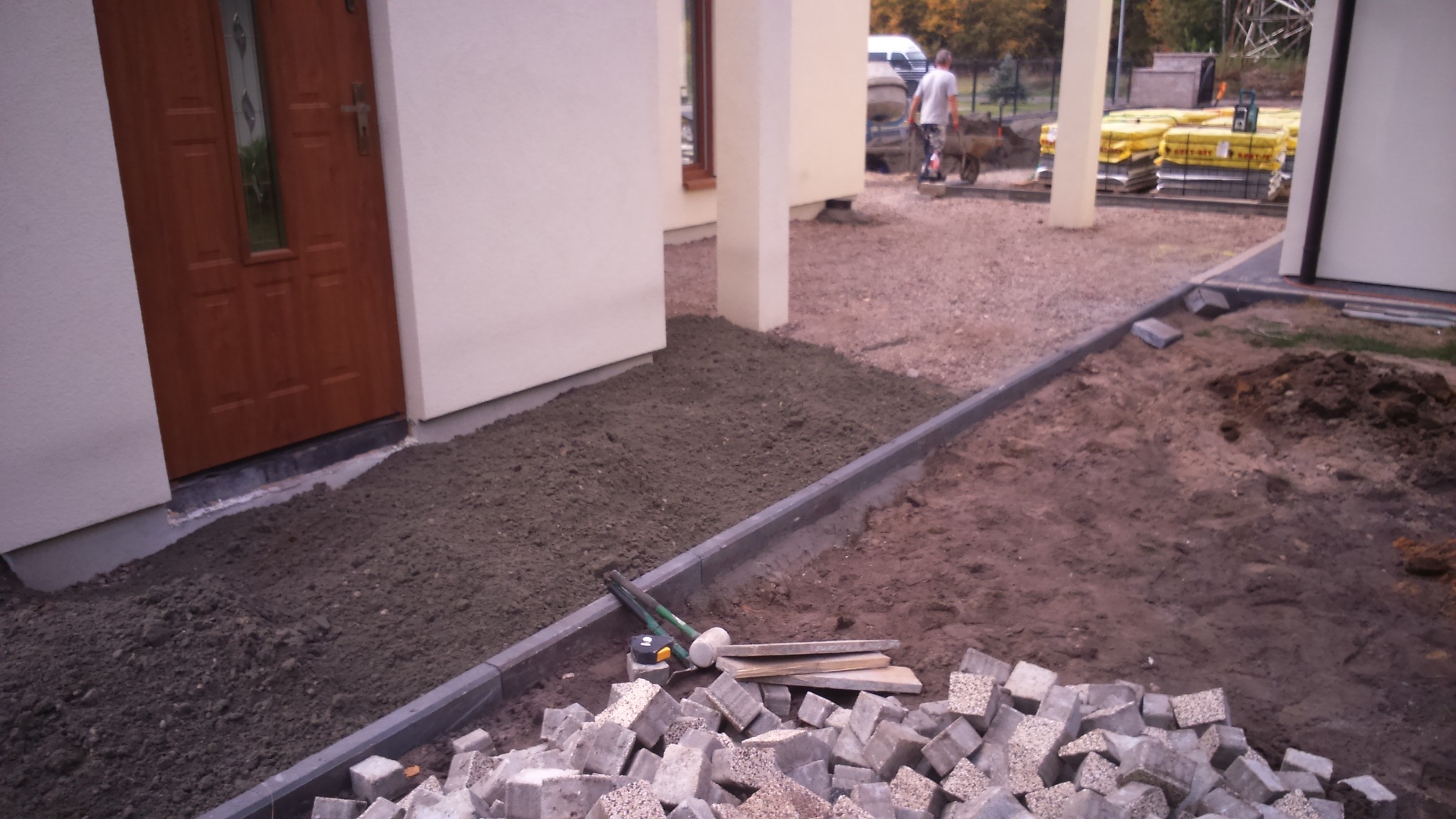 Przygotowanie podłoża pod kostkę brukową przed domem z brązowymi drzwiami, widoczne obrzeża, narzędzia i sterta kostki brukowej.