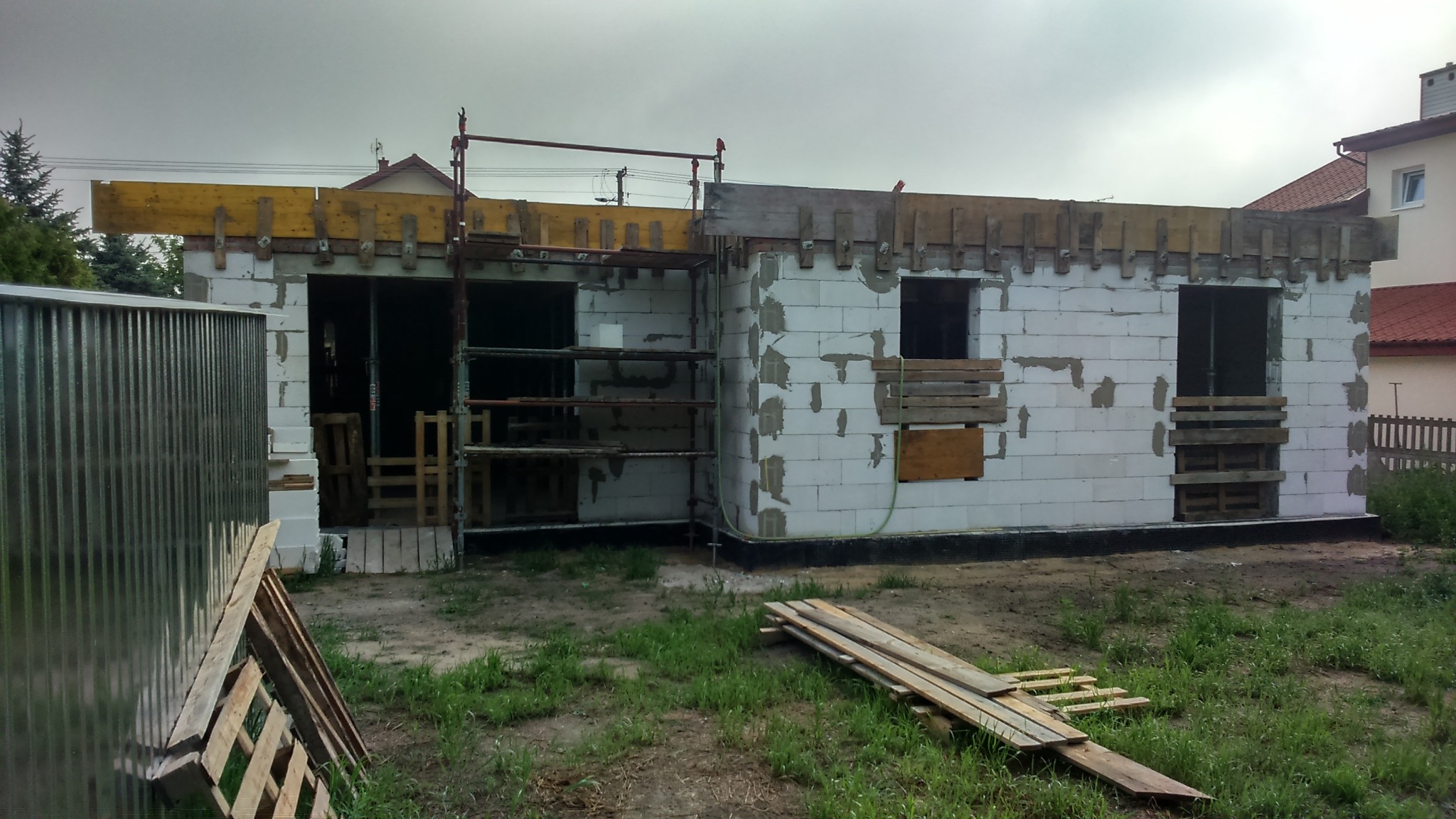 Budowa domu jednorodzinnego w stanie surowym otwartym, ściany z betonu komórkowego, widoczne rusztowania, szalunki na stropie, zabezpieczone otwory okienne deskami.