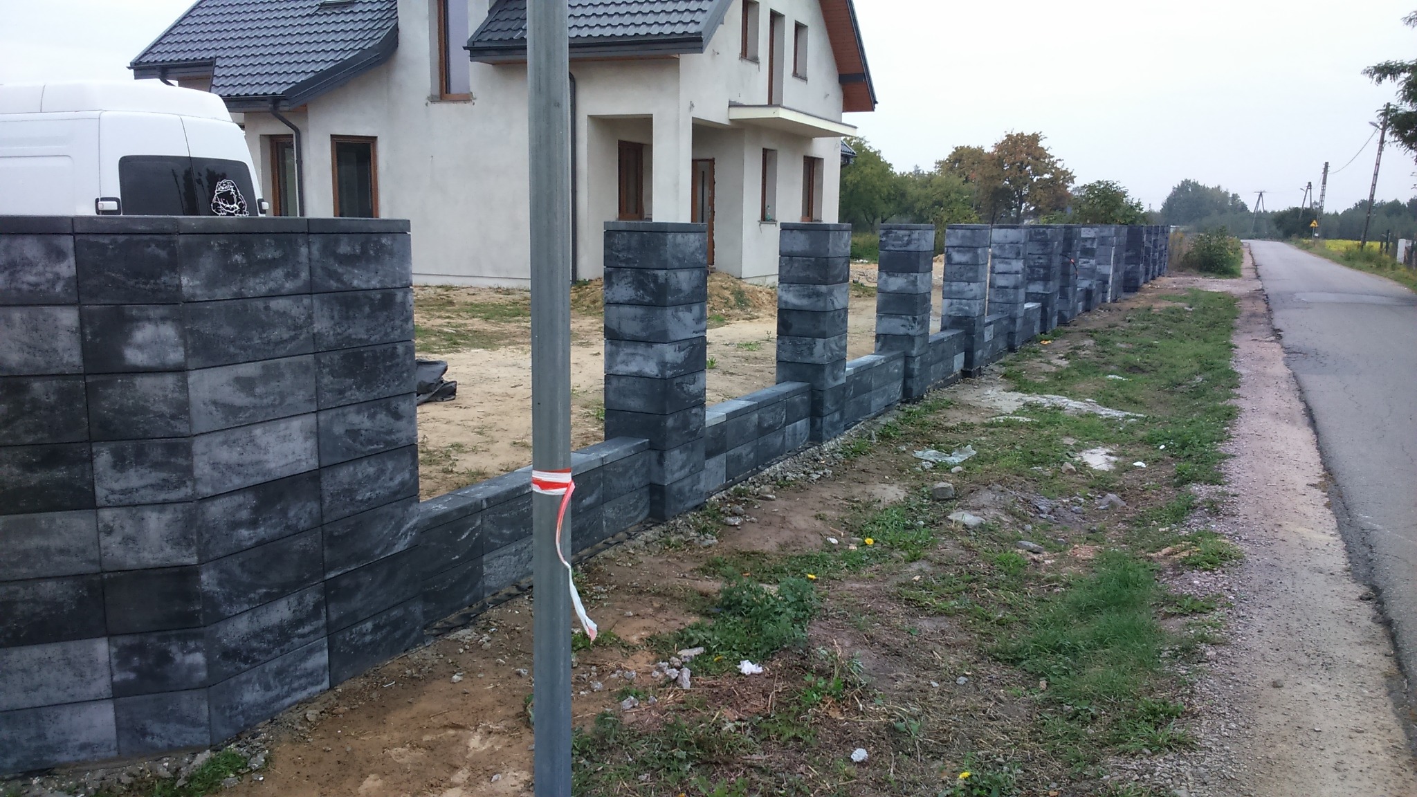 Nowoczesny mur z szarych bloczków betonowych w Piotrkowie Trybunalskim, częściowo ukończony, z widocznym domem w tle i białą furgonetką z naklejką na szybie.