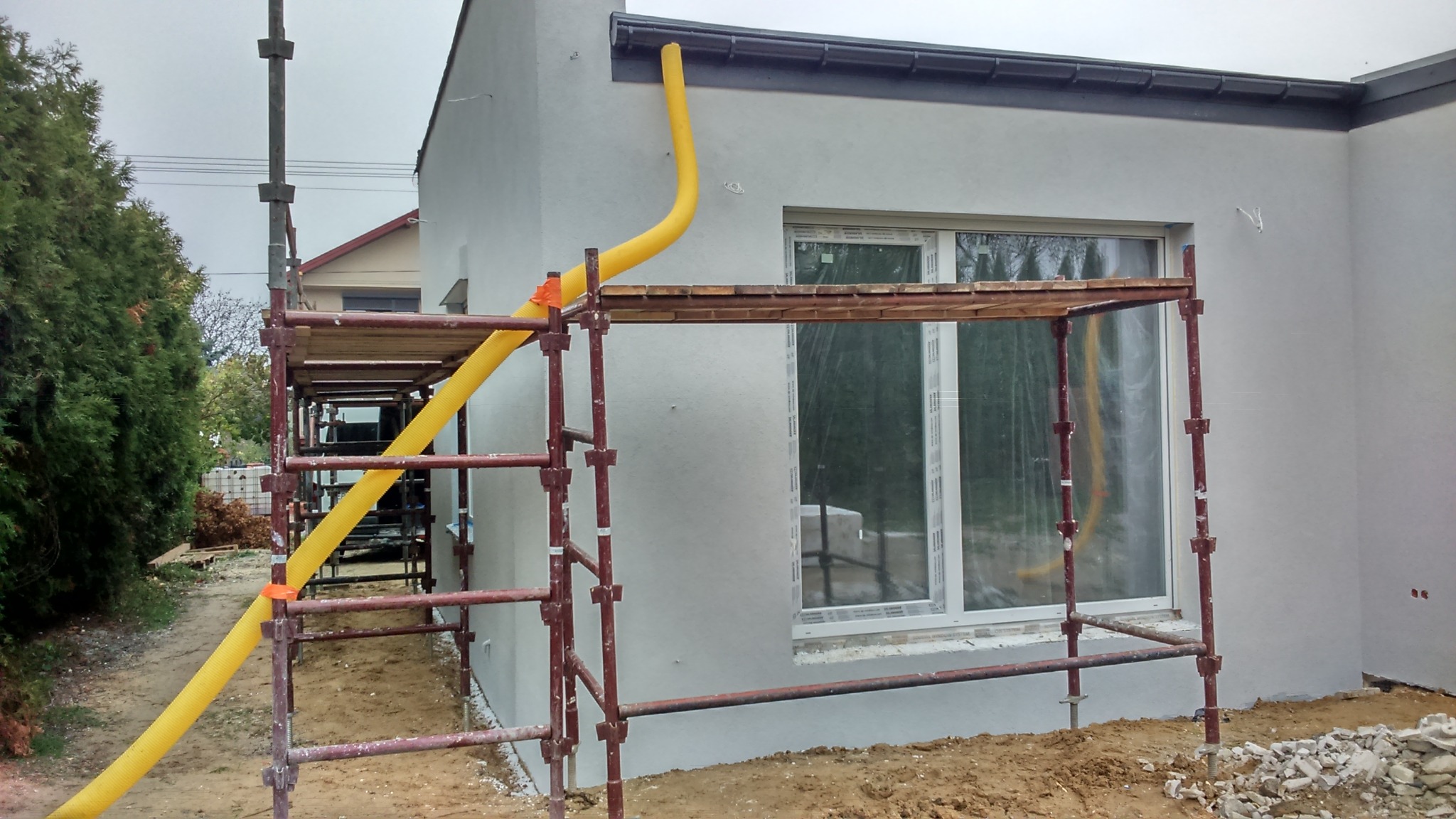 Świeżo otynkowany budynek z oknem PCV, widoczne rusztowanie i żółta rura spustowa do gruzu, tło częściowo zarośnięte zielenią.