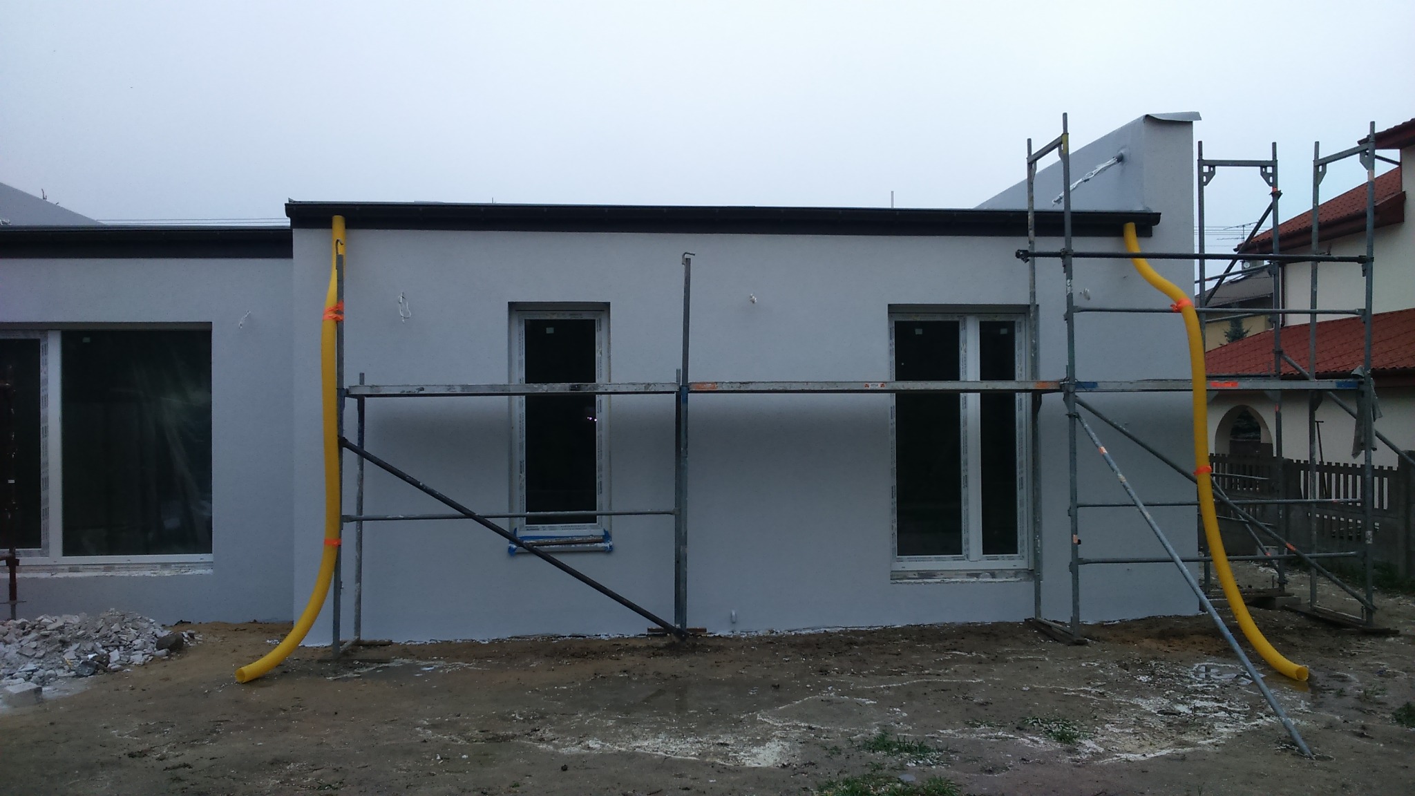 Świeżo otynkowany budynek w trakcie prac wykończeniowych, z widocznymi rusztowaniami i żółtymi rurami spustowymi do gruzu.