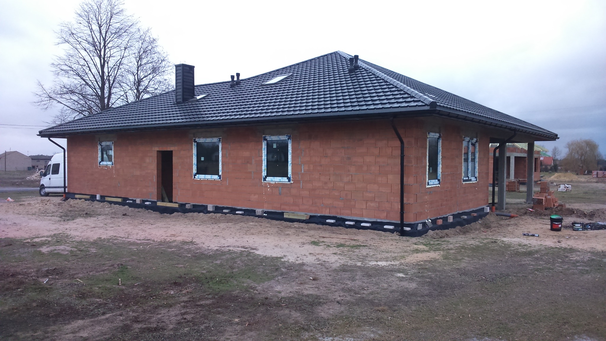 Surowy stan domu jednorodzinnego z czerwonej cegły, z zamontowanymi oknami PCV i czarną dachówką, widoczne przygotowanie do ocieplenia fundamentów, na zewnątrz zaparkowany biały bus.