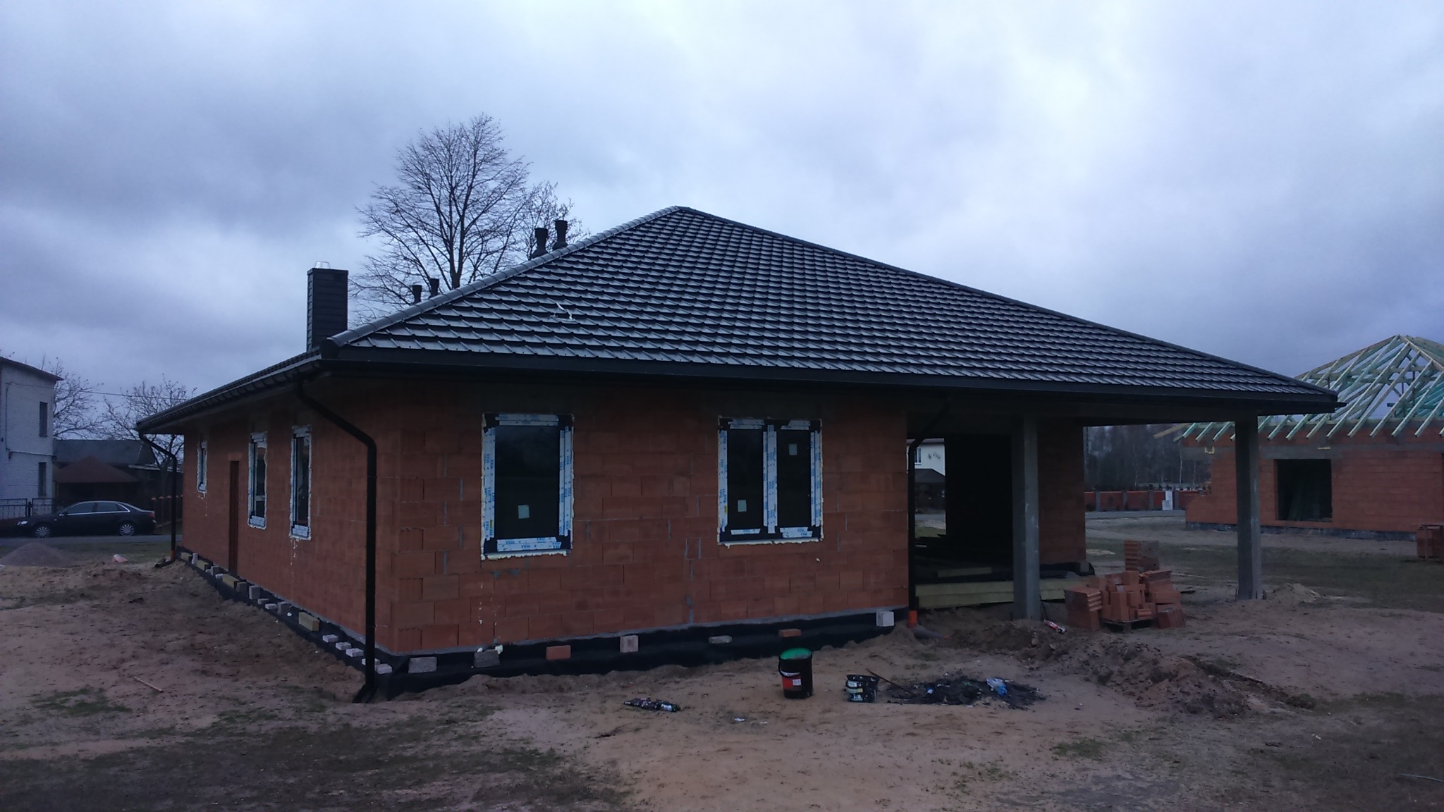 Surowy, jednopiętrowy budynek z czerwonej cegły z zamontowanymi oknami PCV i ciemnym dachem z dachówki, widoczne fundamenty oraz drugi budynek w tle w trakcie budowy.