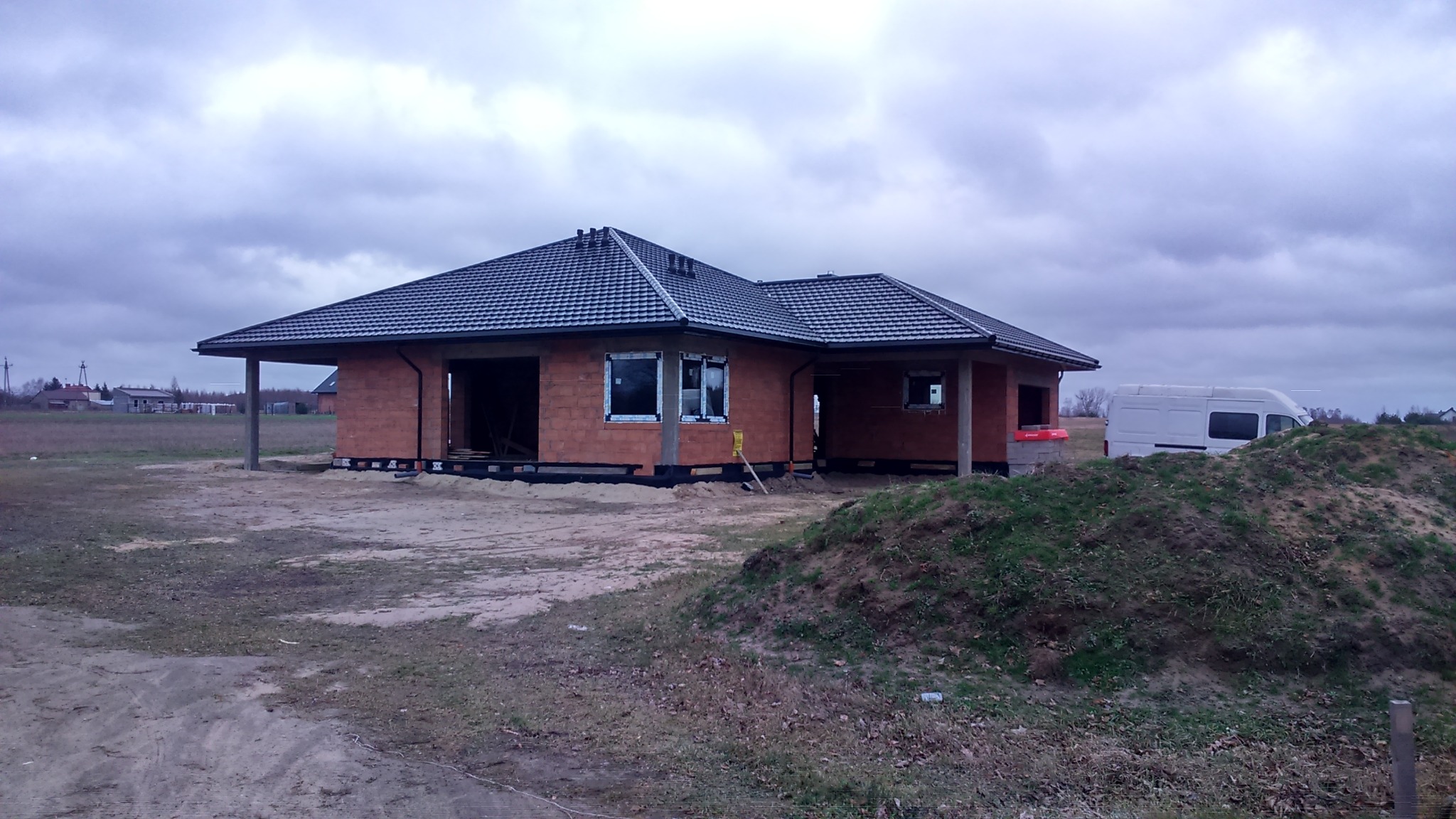 Budowa parterowego domu jednorodzinnego z czerwonej cegły, z czarną dachówką, widoczne okna w białych ramach, na placu budowy stoi biały samochód dostawczy, pochmurne niebo.