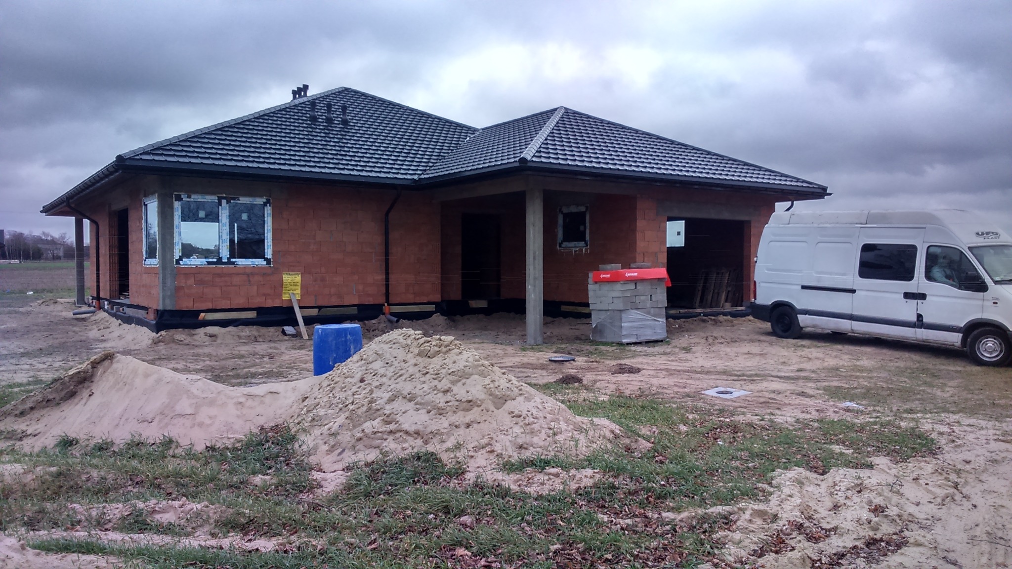Surowy stan domu jednorodzinnego z czerwonej cegły z zamontowanymi oknami PCV, stertą piasku i zaparkowanym busem na placu budowy w pochmurny dzień.