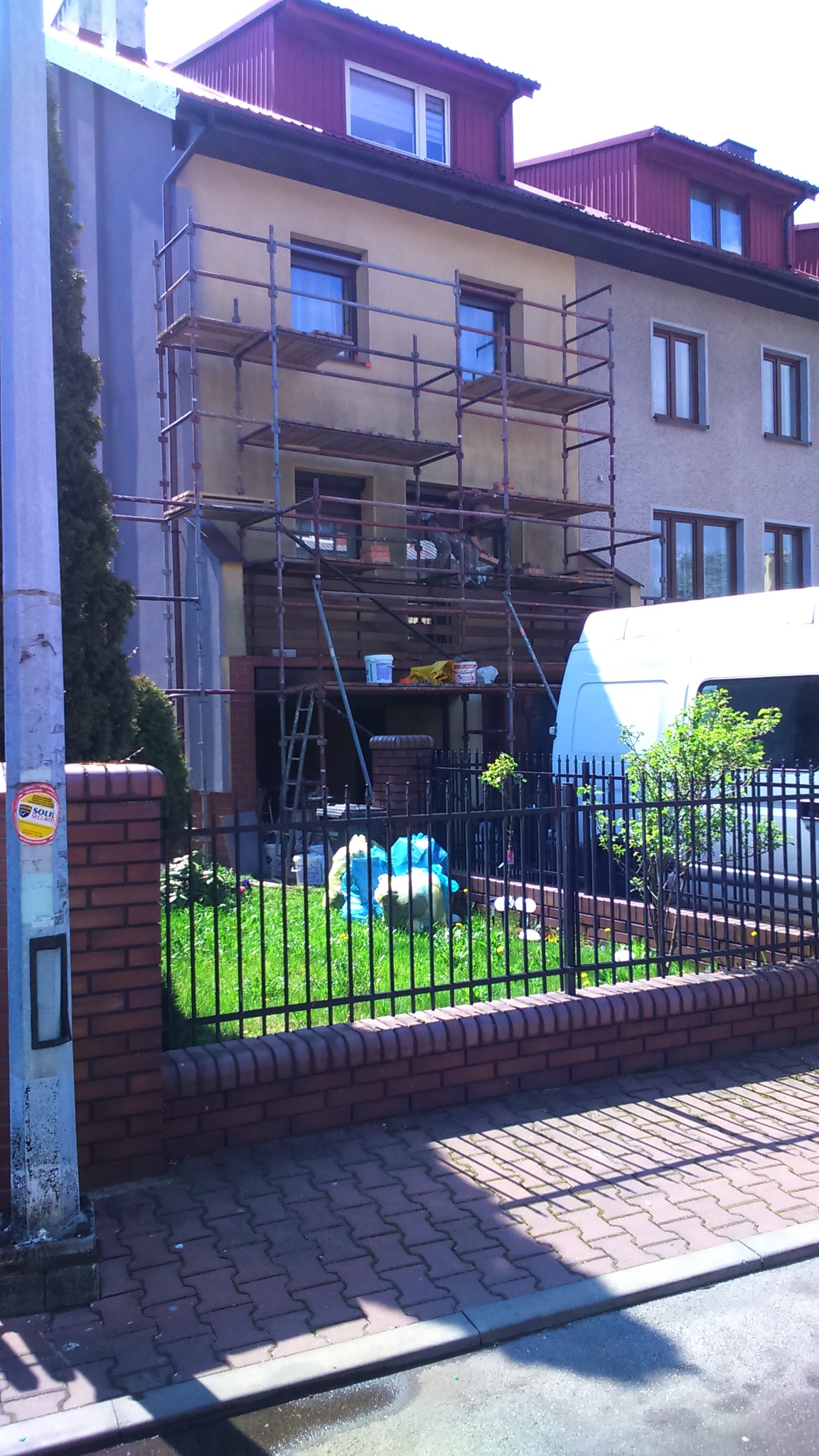 Dwupiętrowy dom w trakcie renowacji elewacji, z ustawionymi rusztowaniami, widoczny zza niskiego murku i metalowego ogrodzenia.