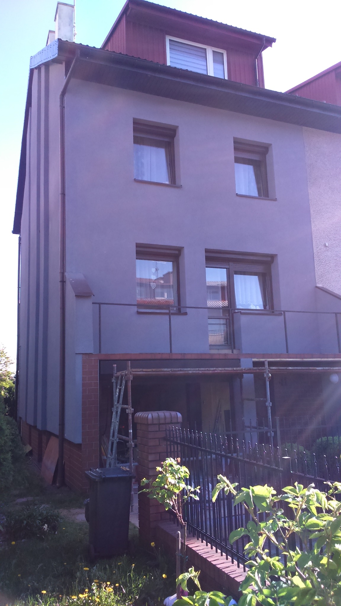 Ocieplony dom z szarą elewacją, widoczne rusztowania i drabina wskazujące na trwające prace wykończeniowe, z elementami ceglanymi u podstawy.