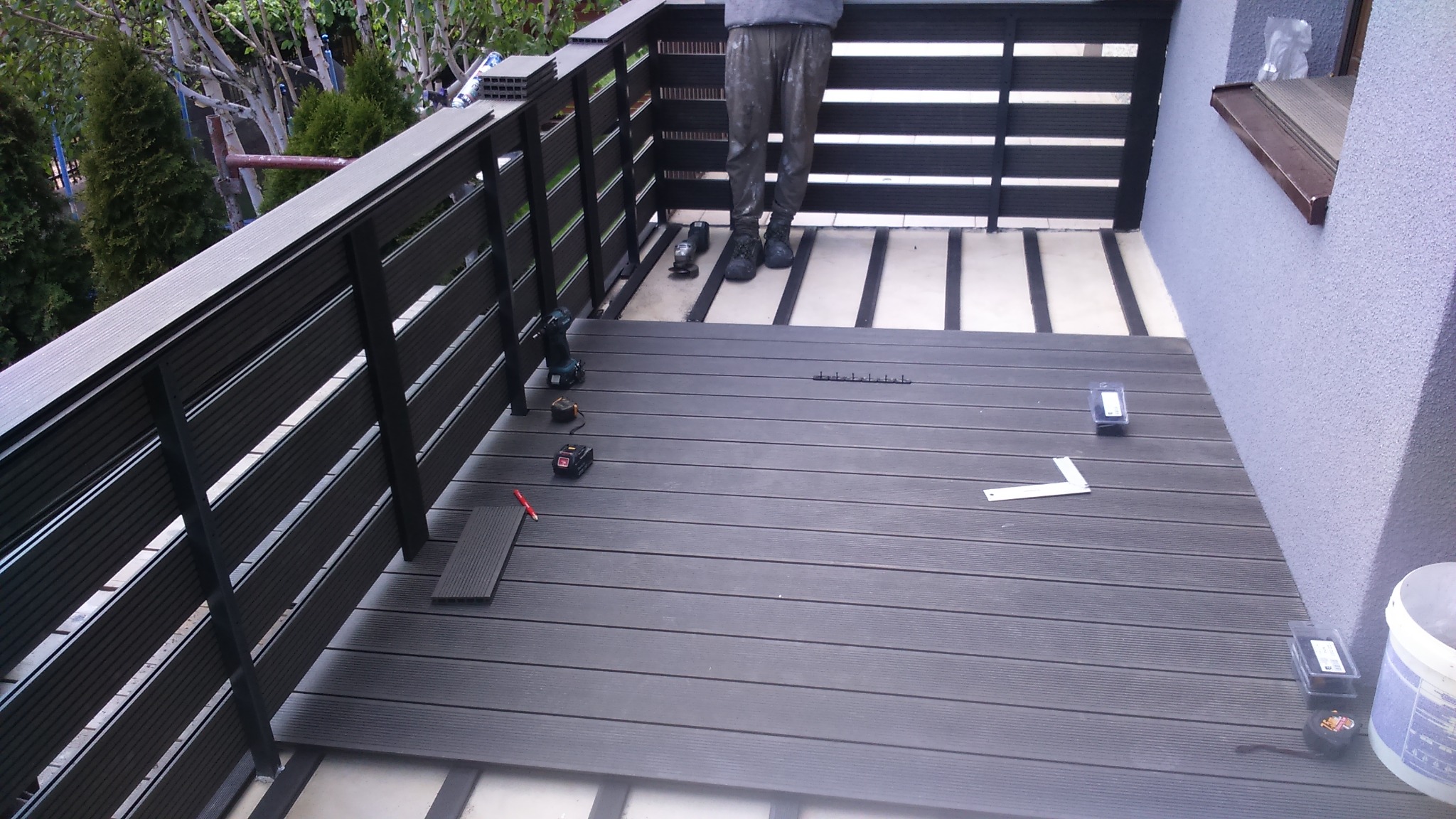 Montaż desek kompozytowych na tarasie: widoczne deski, fragment balustrady z poziomych listew, narzędzia pracy (wiertarka, miara, poziomica), oraz częściowo widoczna osoba w roboczym ubraniu.