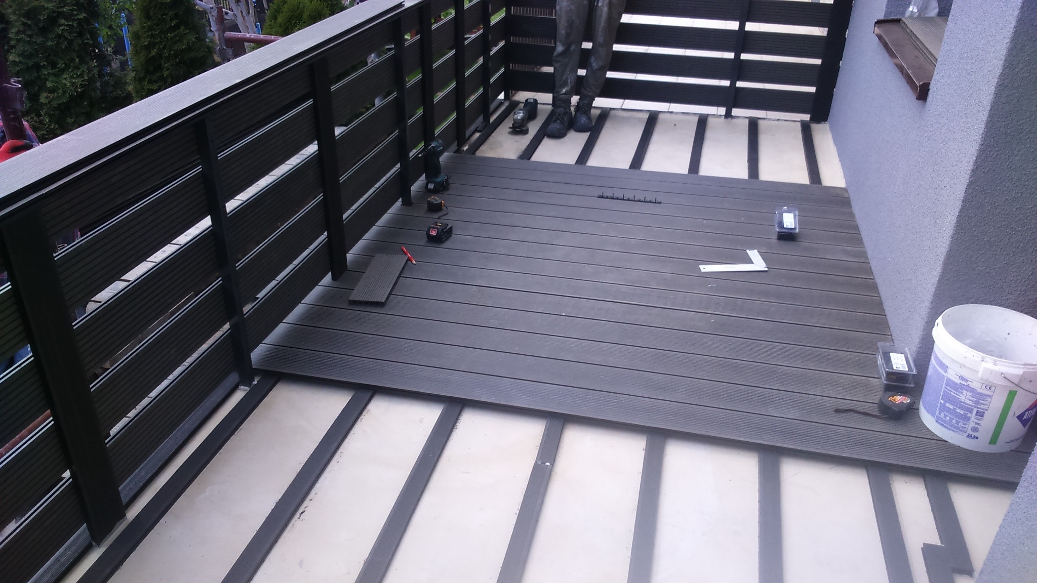 Montaż desek kompozytowych na tarasie, widoczne narzędzia pracy, częściowo wykończona powierzchnia, czarne barierki balkonu.