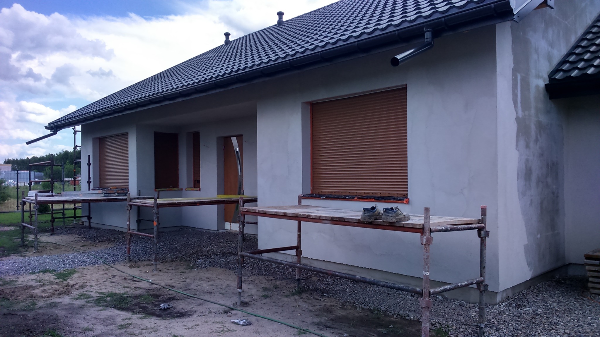 Świeżo otynkowany dom jednorodzinny z szarą elewacją, brązowymi roletami zewnętrznymi, czarnym dachem i rozstawionymi rusztowaniami, na których leżą robocze buty.