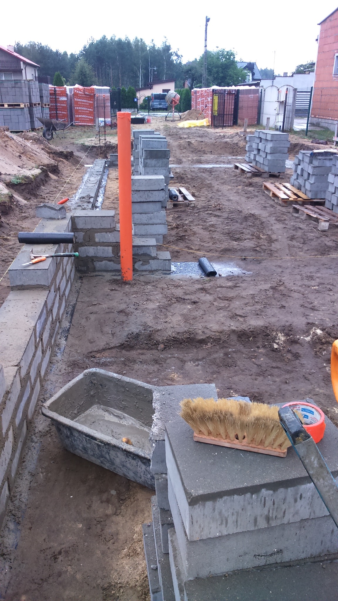 Budowa fundamentów z betonowych bloczków: widoczny wykop, częściowo wymurowane ściany fundamentowe, pomarańczowa rura pionowa, zaprawa w kuwecie, szczotka i taśma miernicza na bloczkach, w tle...