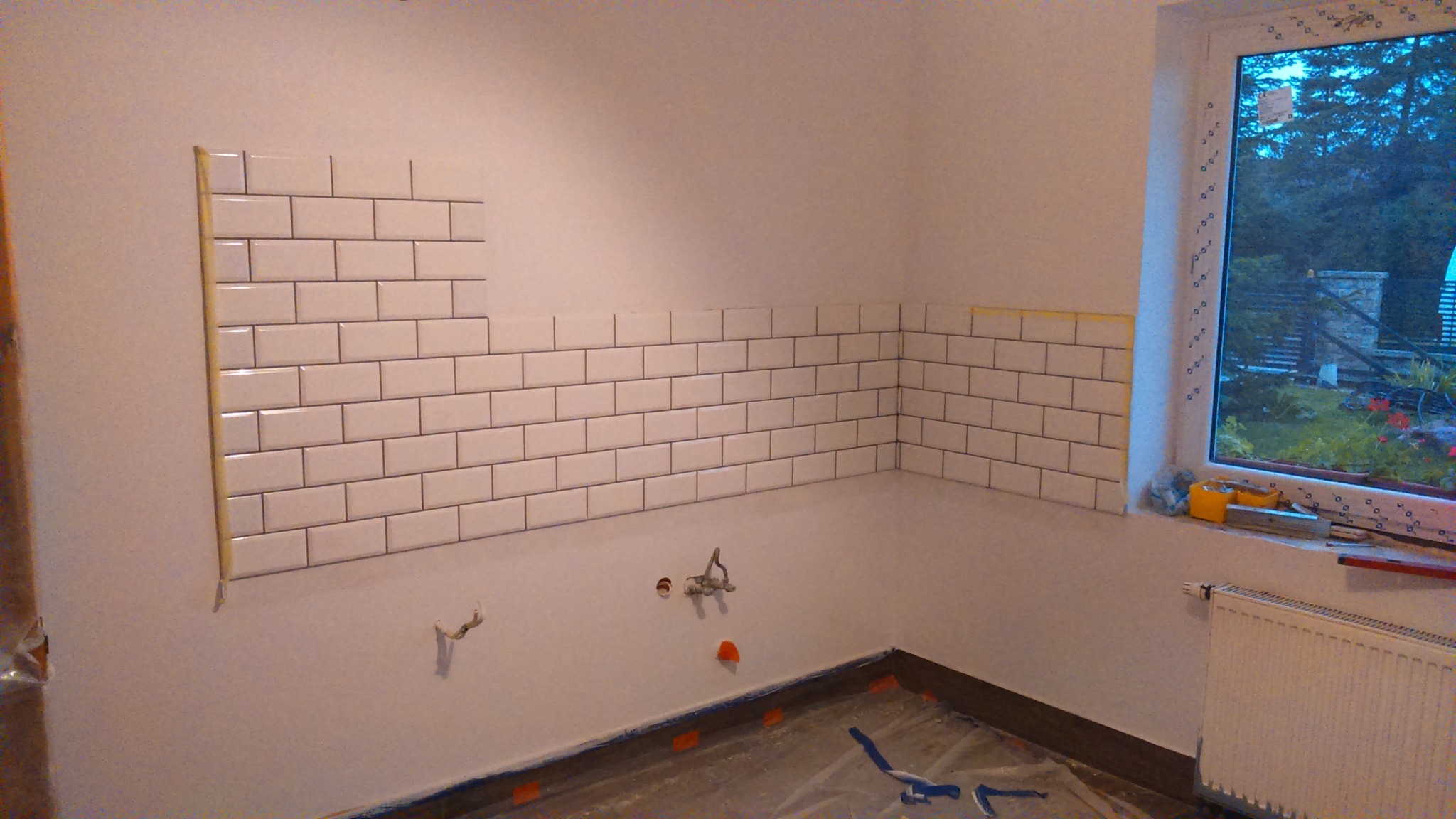 Układanie białych, prostokątnych płytek typu metro na ścianie w kuchni, widoczne fragmenty instalacji wodnej, okno z widokiem na ogród, zabezpieczona folią podłoga i grzejnik.