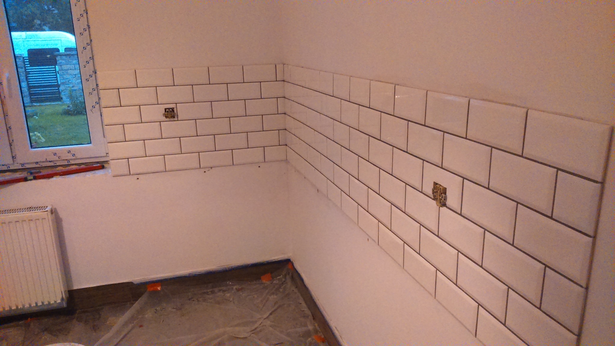 Białe, prostokątne płytki ułożone na ścianie w kuchni, z widocznymi puszkami elektrycznymi i oknem z widokiem na zewnątrz, podłoga zabezpieczona folią malarską.