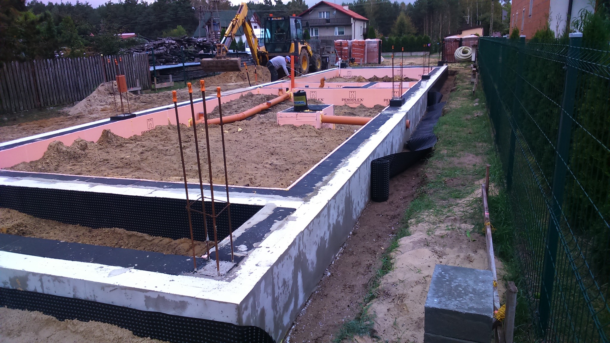 Wykopane fundamenty domu jednorodzinnego z widocznym zbrojeniem, izolacją termiczną w kolorze różowym, drenażem oraz częściowo wylanym betonem, na tle koparki i cegieł.