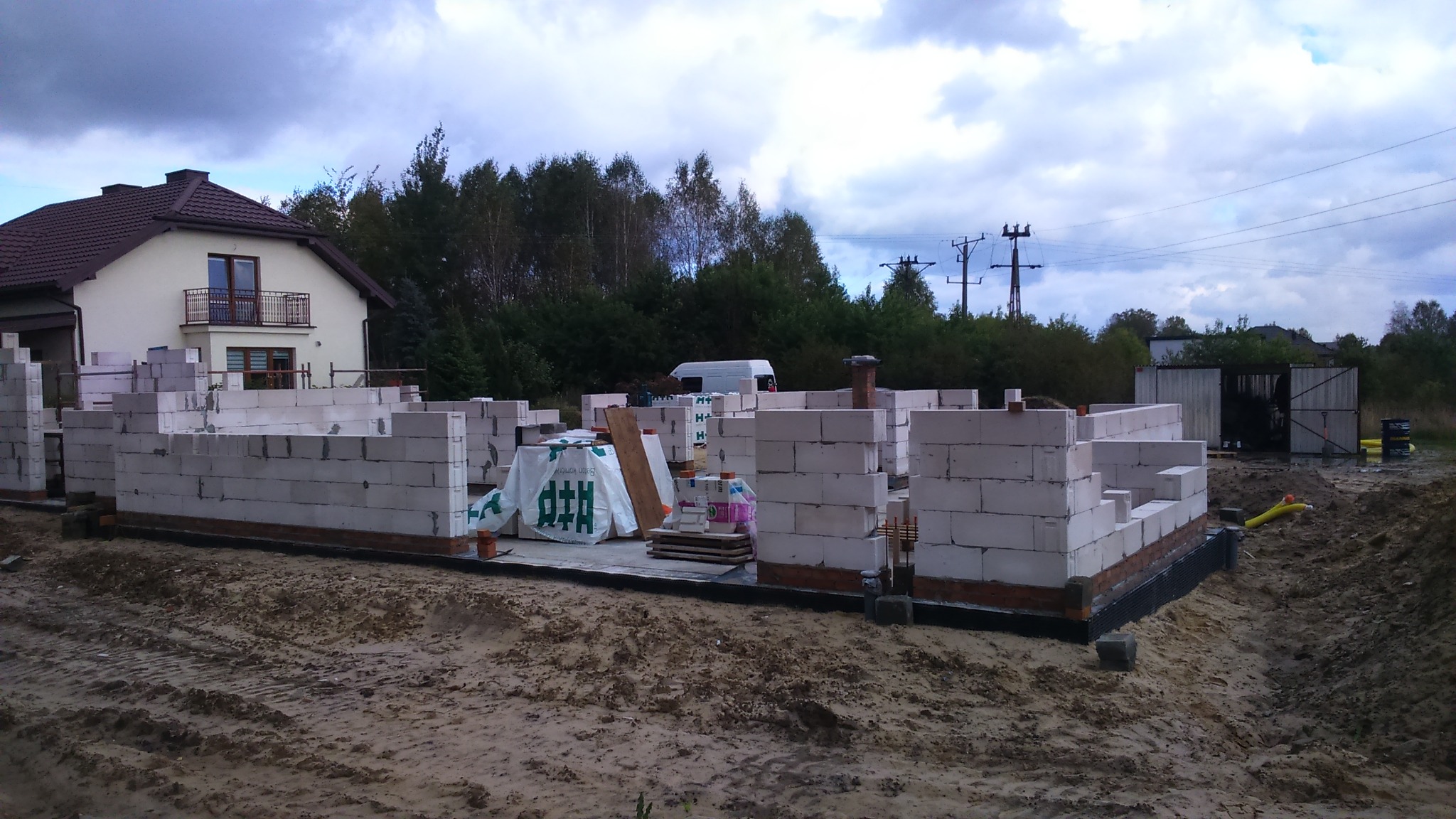 Wznoszenie ścian domu z bloczków betonowych na fundamencie, widoczne materiały budowlane, w tle dom jednorodzinny i pochmurne niebo.