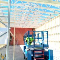 Konstrukcja drewniana budynku w trakcie montażu, widoczny pracownik w kamizelce na podnośniku, błękitne niebo w tle.