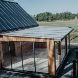 Nowoczesna dobudówka z dużymi przeszkleniami i drewnianą elewacją, widok z góry na taras i otaczający krajobraz.