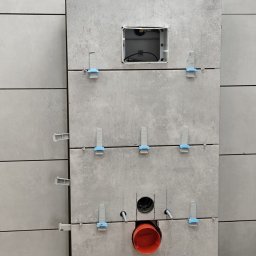 Złota Rączka - Szare płytki imitujące beton, ułożone na ścianie w łazience, z widocznymi systemami poziomowania płytek i otworem na instalację sanitarną.