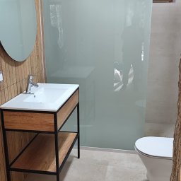 Złota Rączka - Nowoczesna łazienka z umywalką na drewnianym stelażu z czarnymi metalowymi nogami, owalne lustro, kabina prysznicowa z mlecznego szkła i biała toaleta.