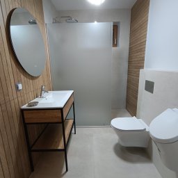 Złota Rączka - Nowoczesna łazienka z drewnianymi akcentami: owalne lustro z podświetleniem nad umywalką z szafką na metalowym stelażu, kabina prysznicowa ze szkła matowego, podwieszana toaleta w minimalistycznym...