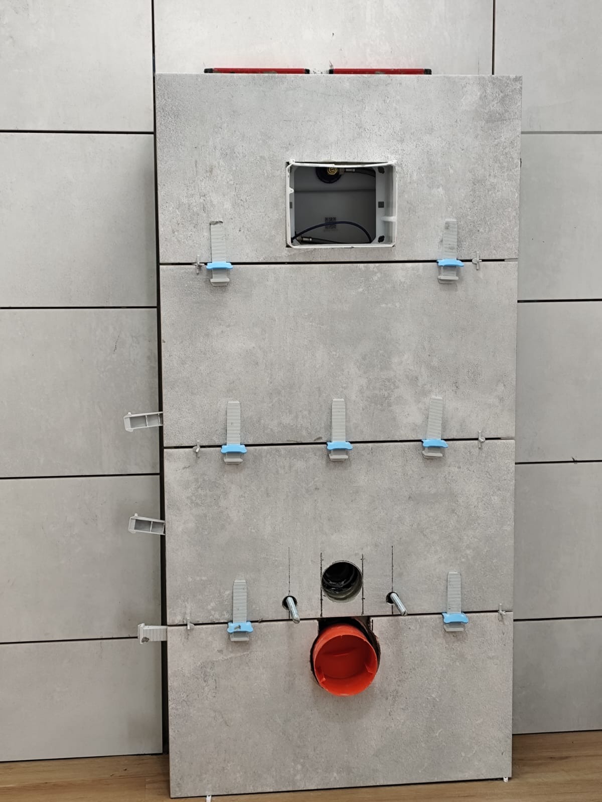 Szare płytki imitujące beton, ułożone na ścianie w łazience, z widocznymi systemami poziomowania płytek i otworem na instalację sanitarną.
