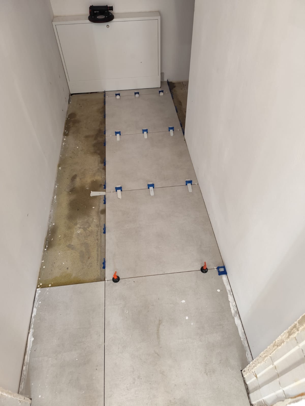 Układanie szarych płytek podłogowych w wąskim korytarzu, widoczne narzędzia do poziomowania i krzyżaki dystansowe, część podłogi jeszcze nie wykończona.