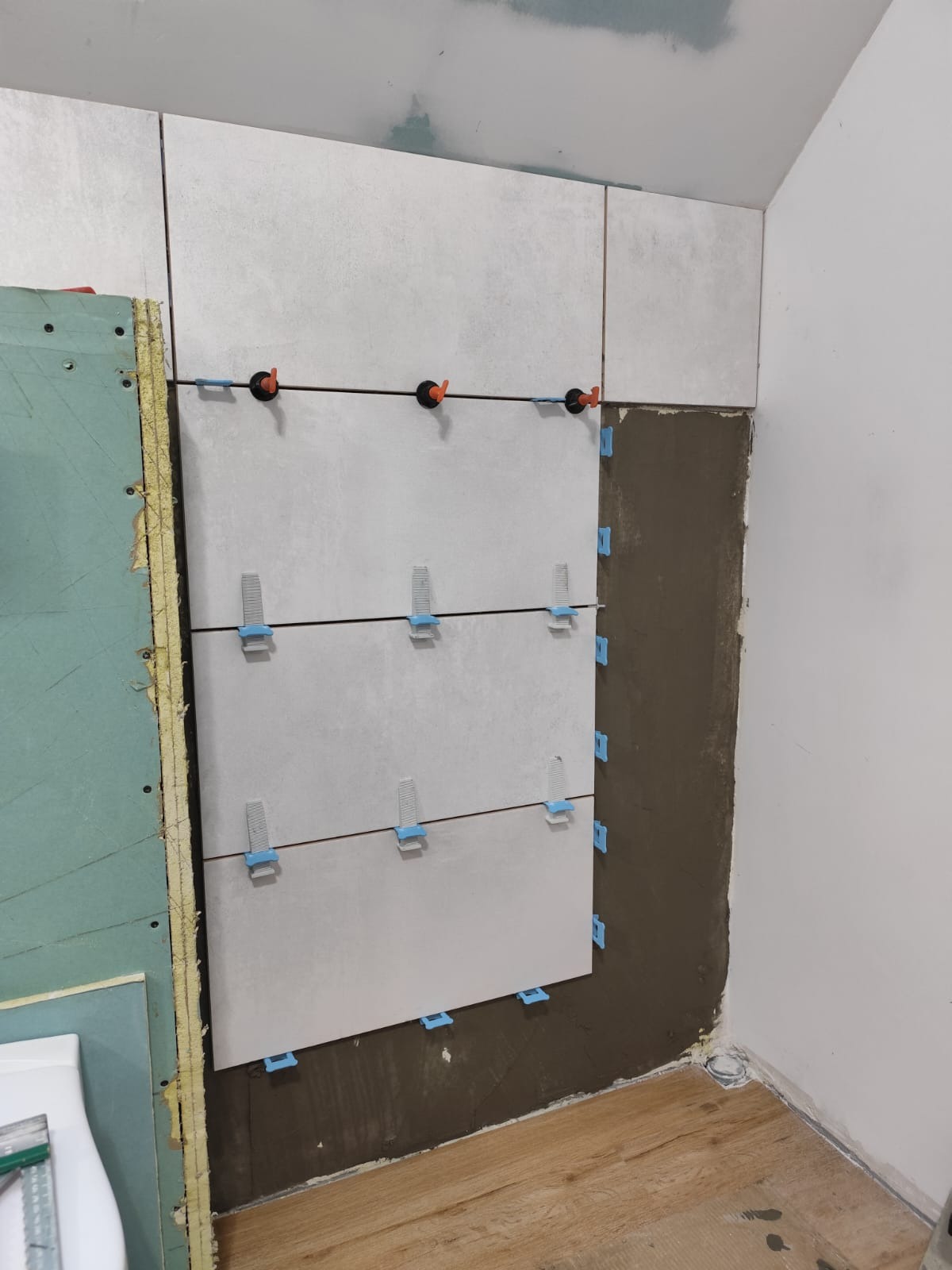 Montaż płytek ceramicznych na ścianie w łazience z użyciem poziomujących klipsów i krzyżyków dystansowych, widoczna surowa ściana z zaprawą i płyta gipsowa po lewej stronie.