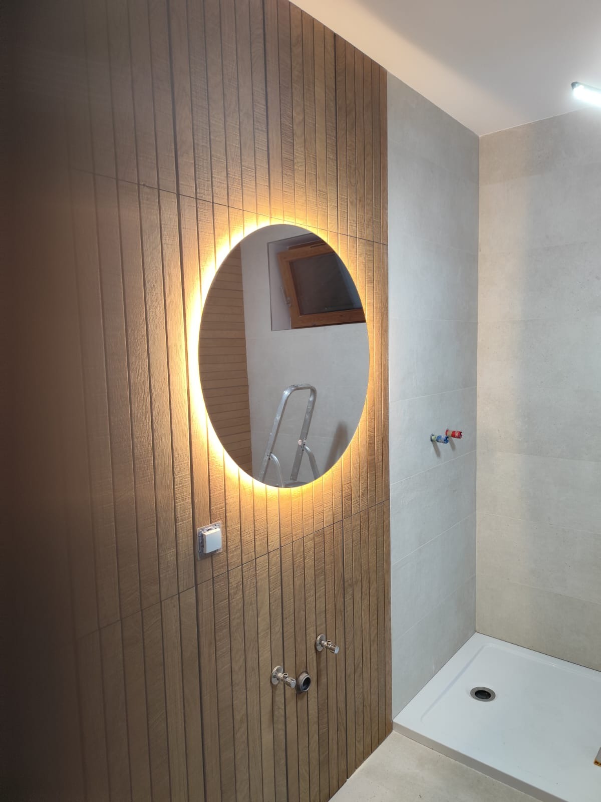 Oświetlone okrągłe lustro na ścianie z drewnopodobnych pionowych paneli w łazience w trakcie wykańczania, widoczna drabina i przyłącza wody.