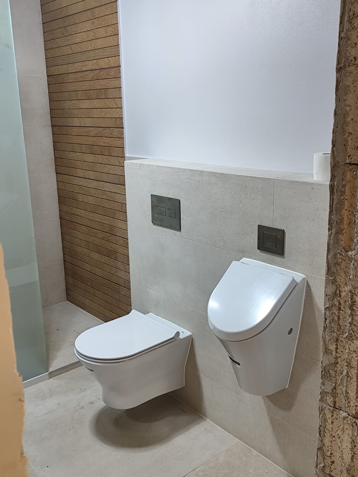 Nowoczesna łazienka z podwieszaną toaletą i bidetem, drewniana okładzina ściany w tle, szara płytka na ścianach i podłodze, minimalistyczne przyciski spłukujące.