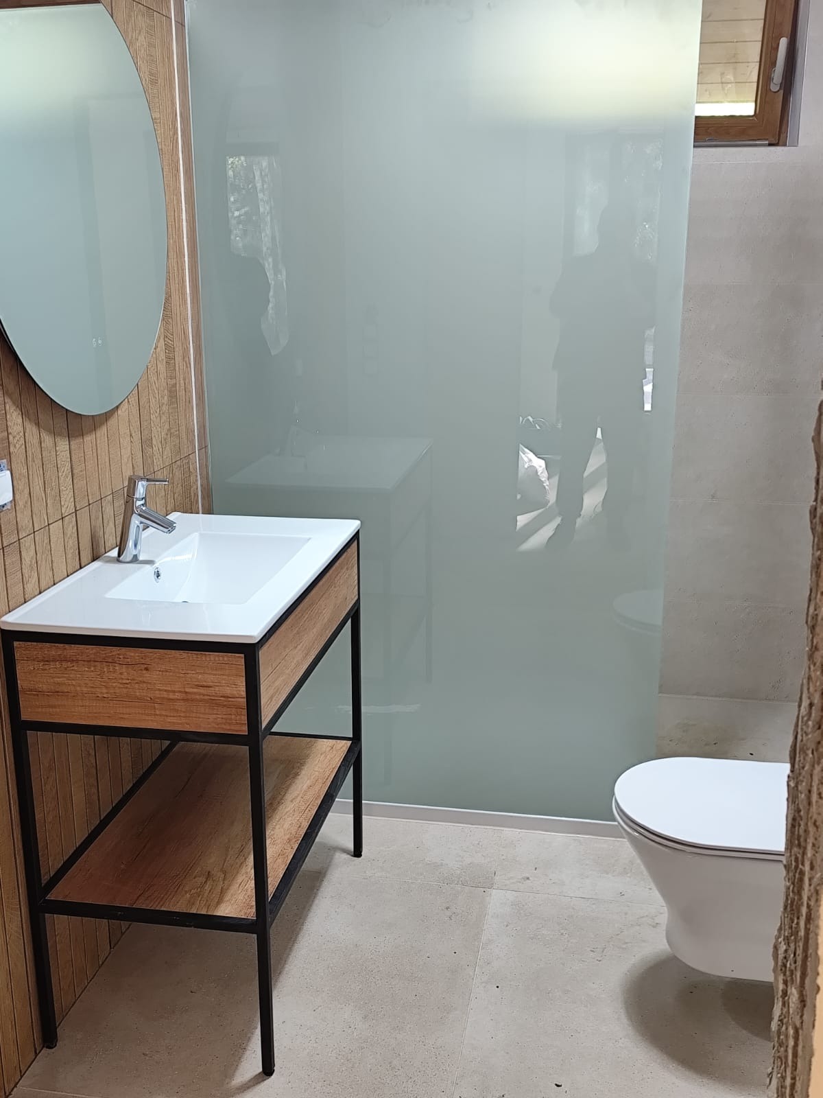 Nowoczesna łazienka z umywalką na drewnianym stelażu z czarnymi metalowymi nogami, owalne lustro, kabina prysznicowa z mlecznego szkła i biała toaleta.