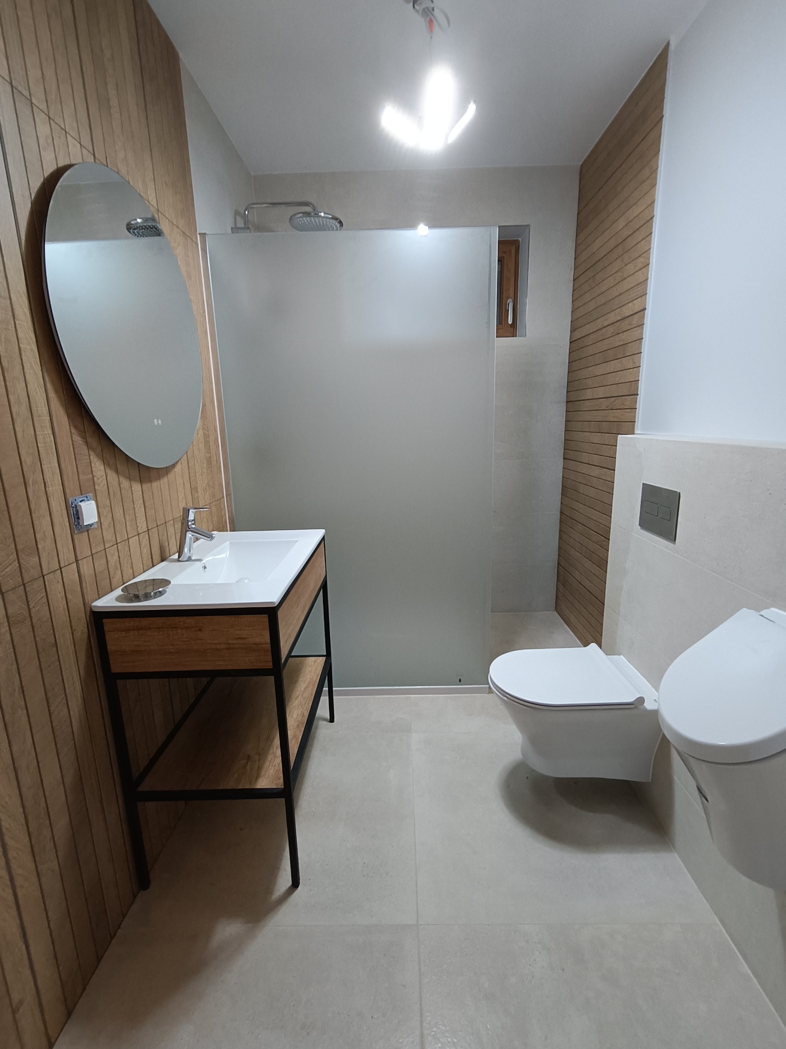 Nowoczesna łazienka z drewnianymi akcentami: owalne lustro z podświetleniem nad umywalką z szafką na metalowym stelażu, kabina prysznicowa ze szkła matowego, podwieszana toaleta w minimalistycznym...