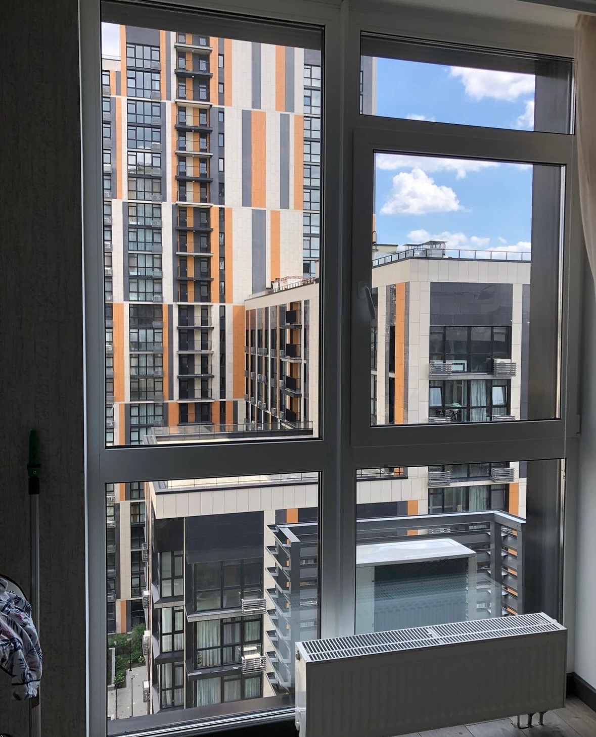 Widok z okna mieszkania na nowoczesne budynki apartamentowe z balkonami w słoneczny dzień, widoczny grzejnik pod oknem.