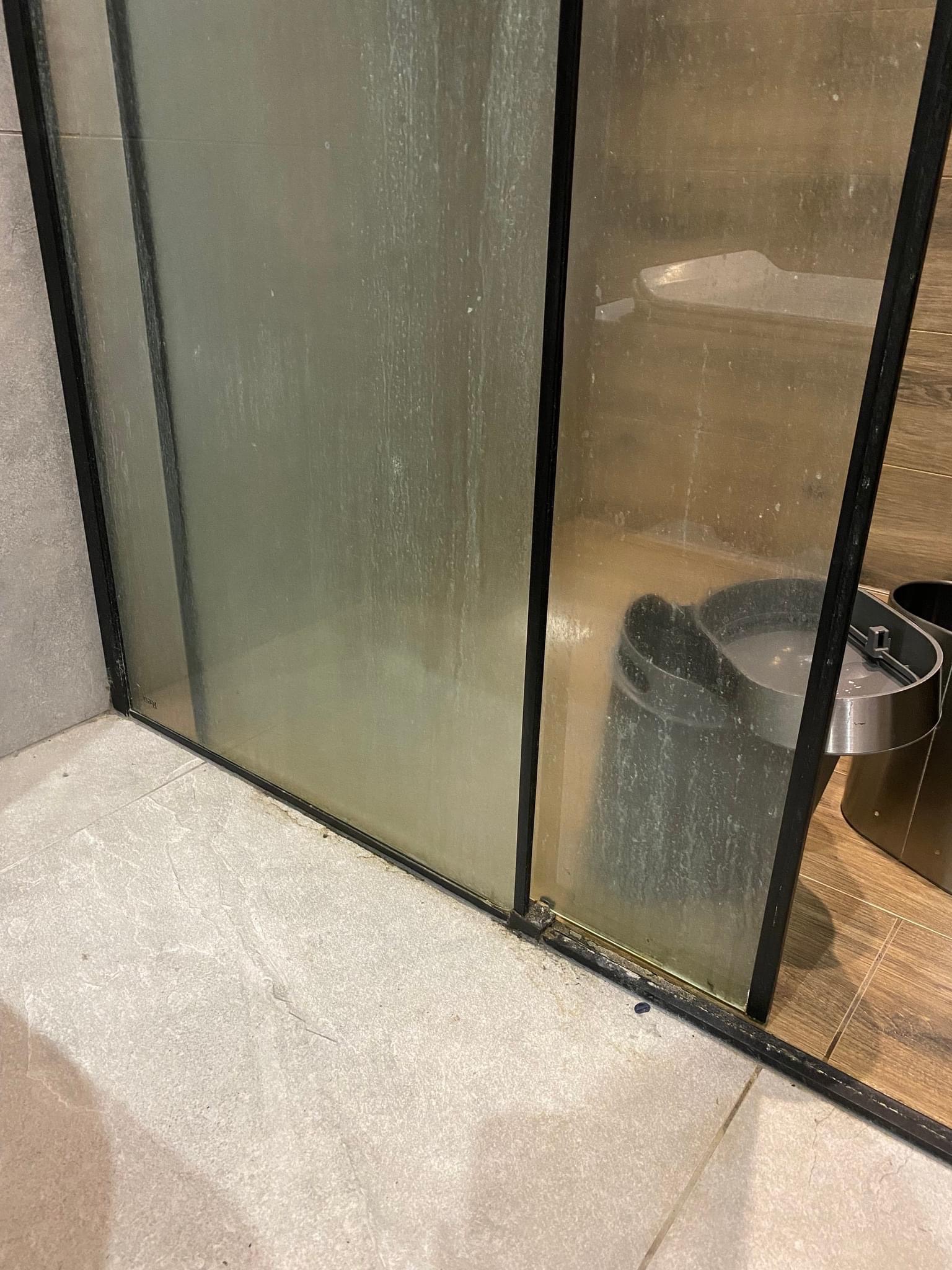 Silnie zabrudzona, matowa szyba kabiny prysznicowej z czarną ramą, widoczne zacieki i osady, fragment podłogi w łazience i kosz na śmieci w tle.