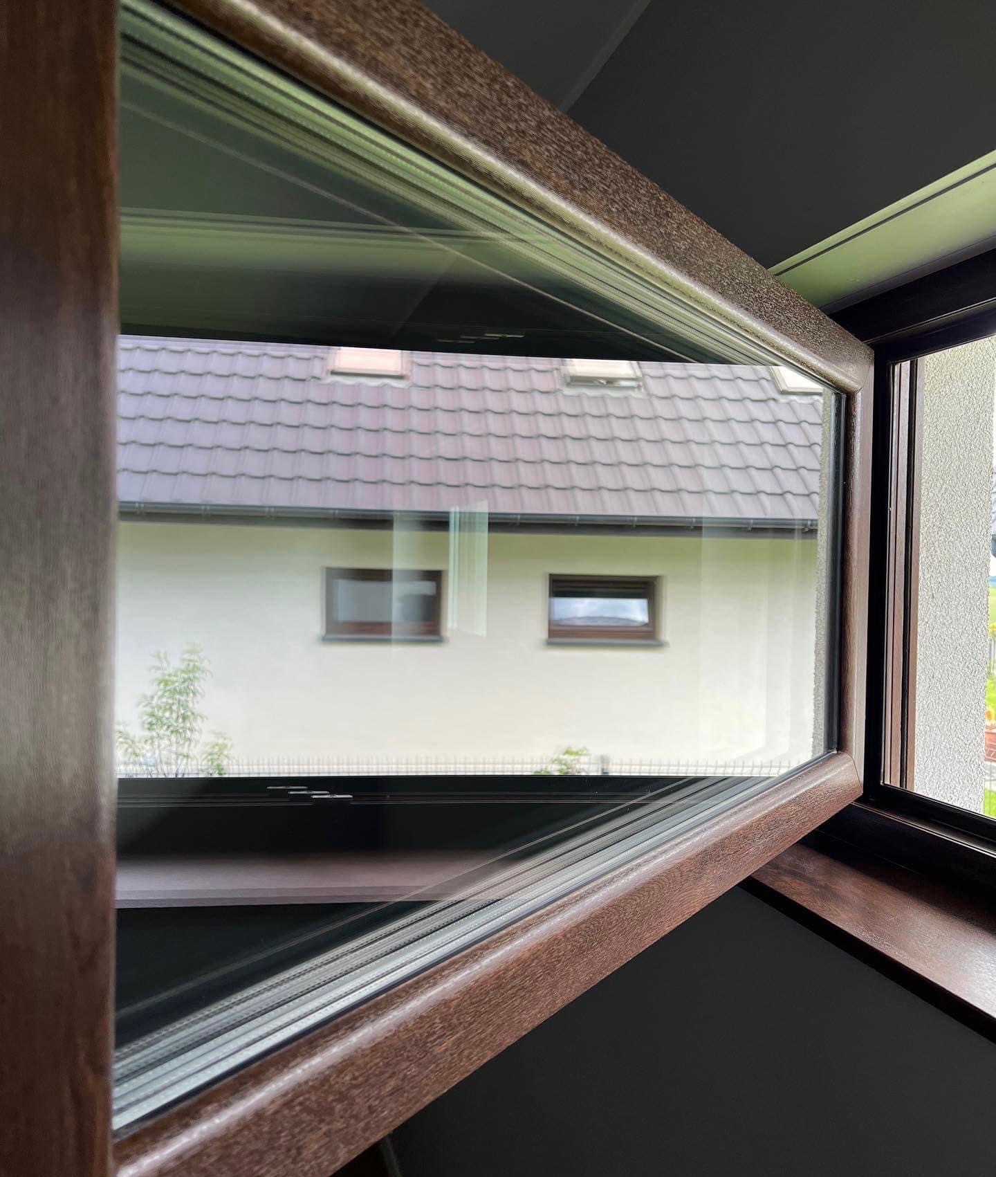 Uchylone okno z brązową ramą, widok na dom z beżową elewacją i brązowym dachem