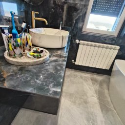 Wykończenie wnętrz i elewacje Machowski Paweł - Nowoczesna łazienka z marmurowymi ścianami, blatem i umywalką, złota armatura, grzejnik panelowy i okno dachowe z roletami.