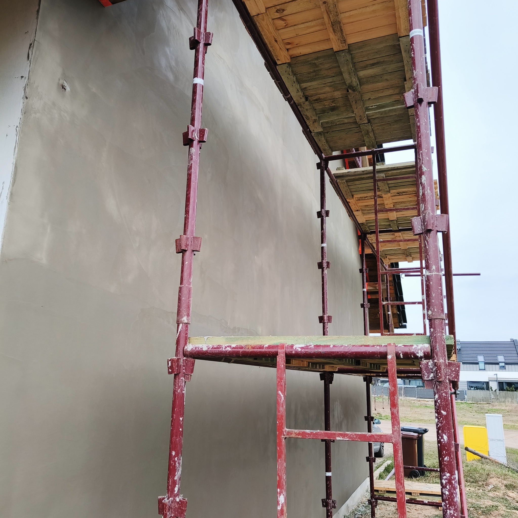 Świeżo nałożona, gładka, szara warstwa tynku na elewacji budynku, widoczna z poziomu rusztowania pomalowanego na czerwono, z fragmentem zabudowy jednorodzinnej w tle.