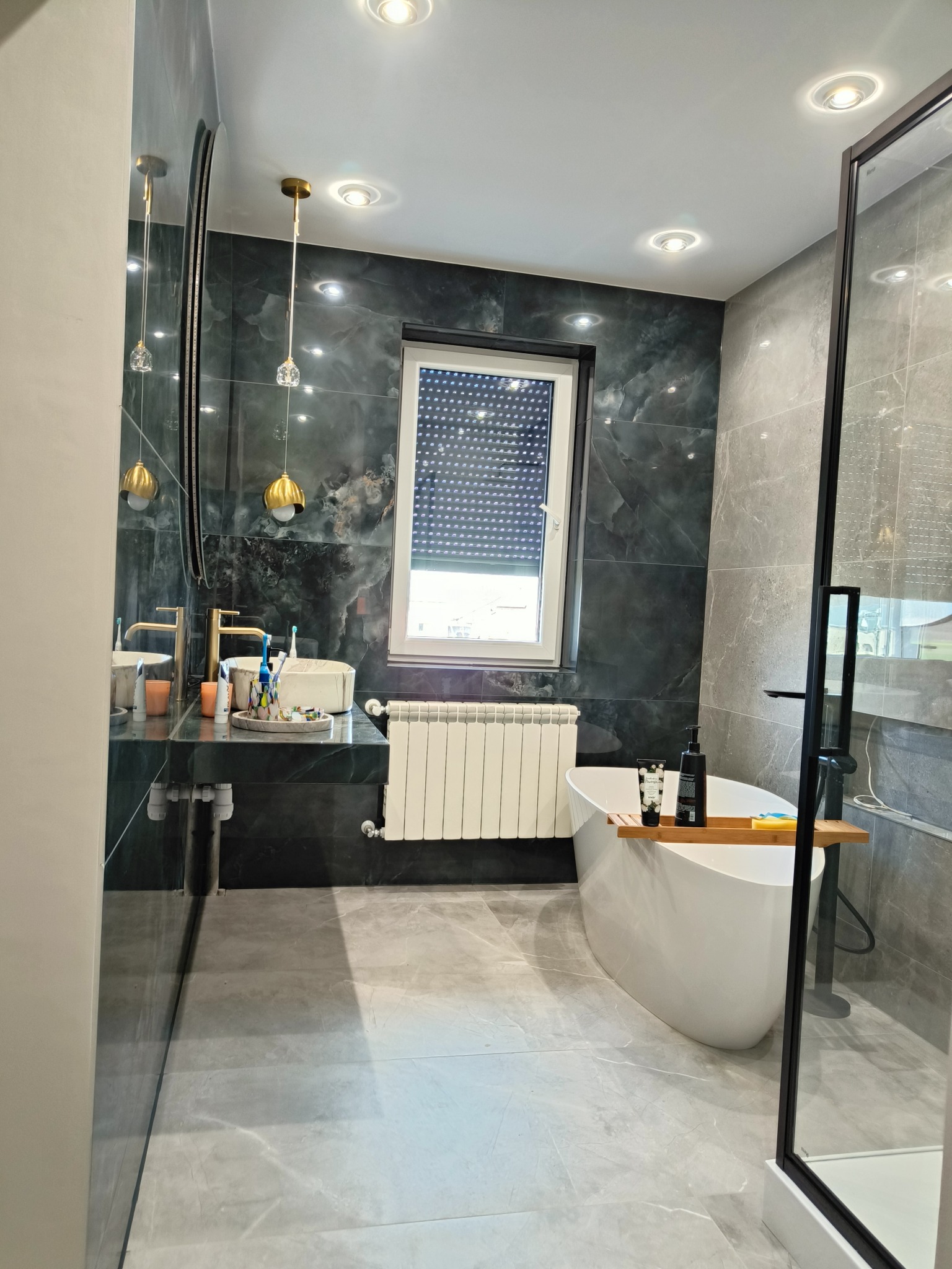 Nowoczesna łazienka z ciemnozielonymi ścianami imitującymi marmur, białą wolnostojącą wanną, przeszkloną kabiną prysznicową i umywalką na ciemnym blacie, oświetlona punktowymi lampami i złotymi...