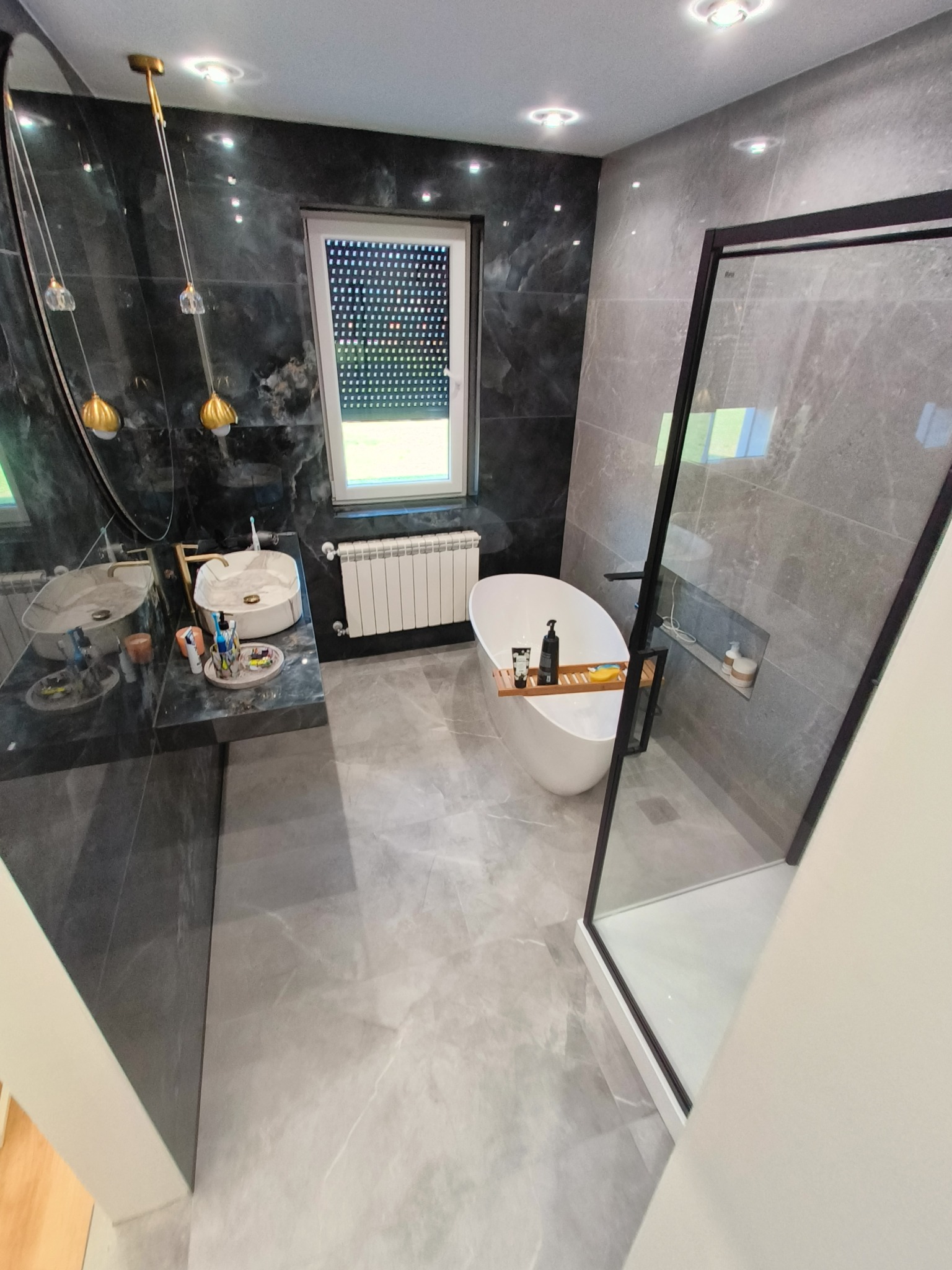 Nowoczesna łazienka z szarymi płytkami imitującymi marmur, wolnostojąca wanna z drewnianą półką na kosmetyki, przeszklona kabina prysznicowa z czarną ramą oraz umywalka nablatowa z baterią...