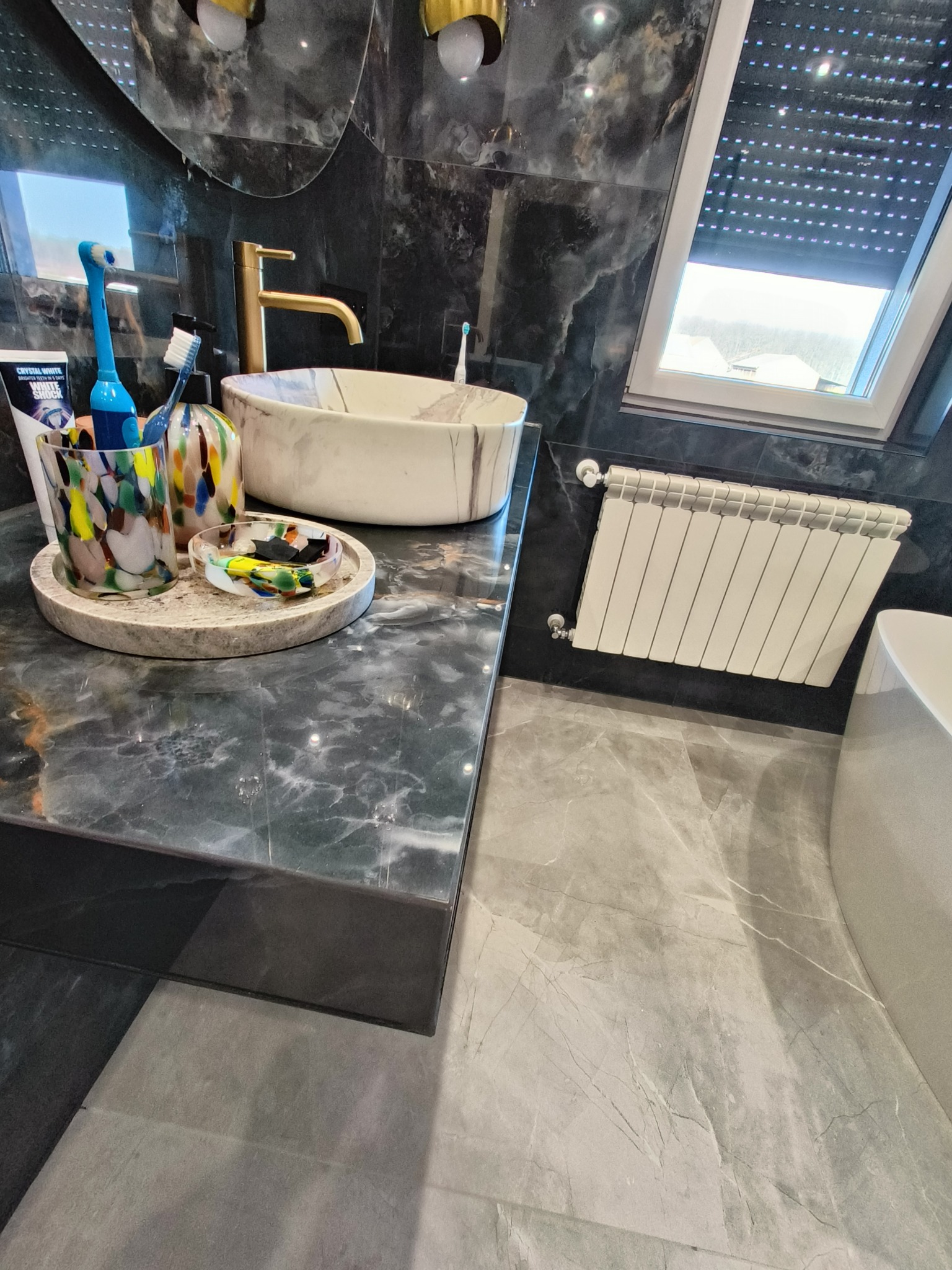 Nowoczesna łazienka z marmurowymi ścianami, blatem i umywalką, złota armatura, grzejnik panelowy i okno dachowe z roletami.