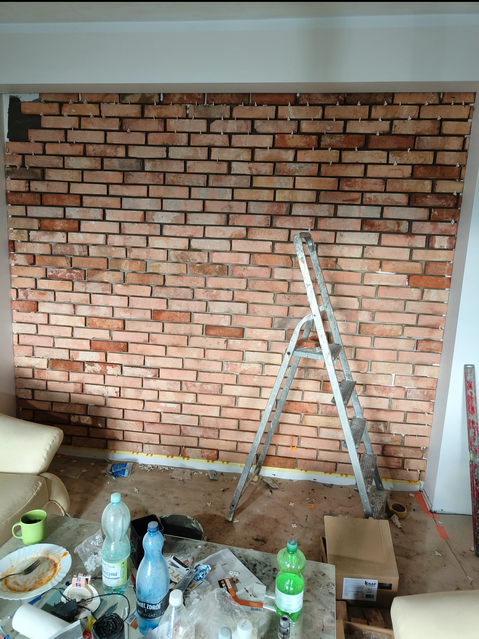 Ściana z cegieł klinkierowych w trakcie montażu, widoczne krzyżyki dystansowe, drabina aluminiowa i bałagan na podłodze.
