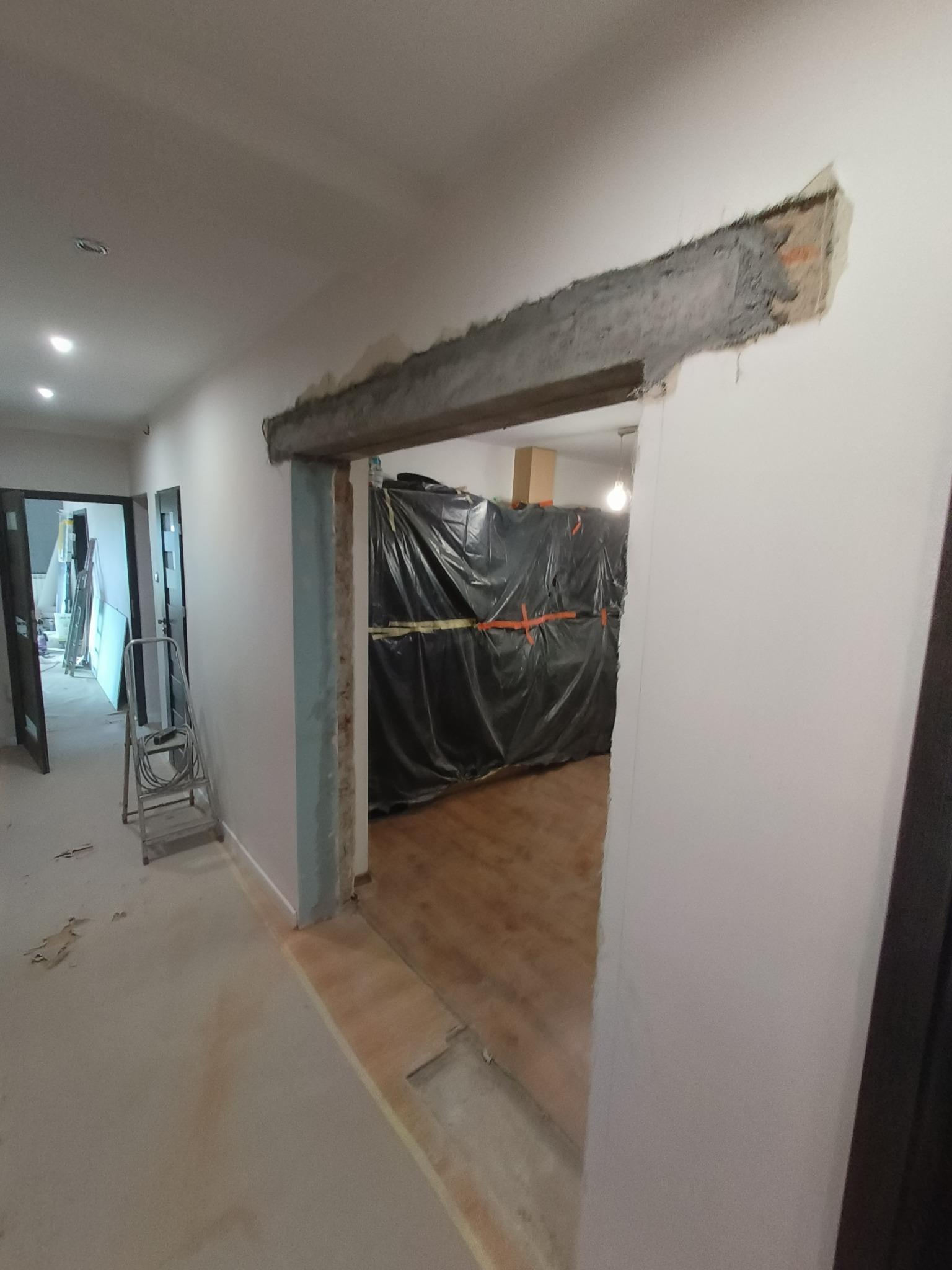 Wnętrze w trakcie remontu: widoczny wycięty otwór w ścianie z surowym betonem, zabezpieczone folią pomieszczenie w tle, drabina aluminiowa i ślady po pracach na podłodze.