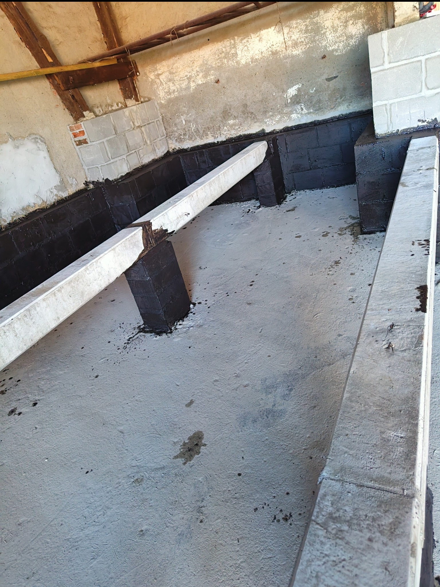Hydroizolacja fundamentów w piwnicy, z widocznymi betonowymi belkami i fragmentami muru z cegieł i pustaków.