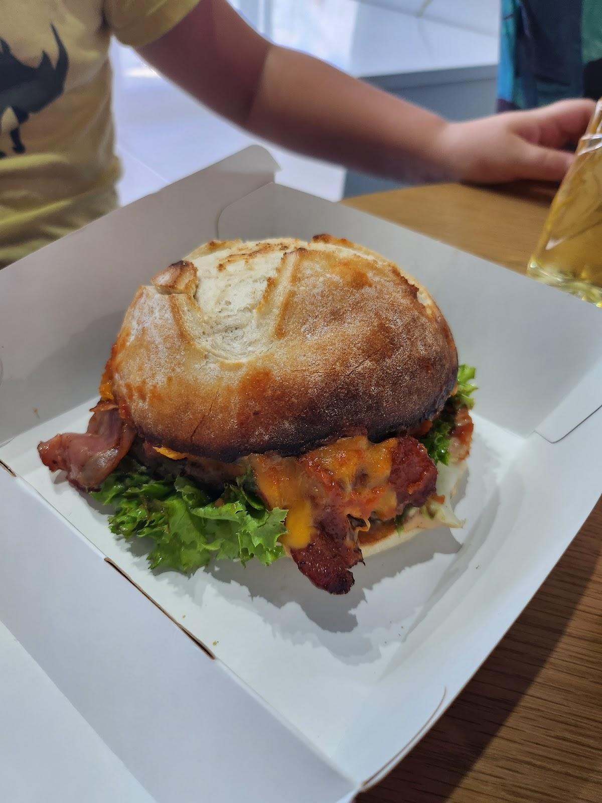 Burger z wołowiną, bekonem, serem i sałatą w białym kartonowym pudełku, widoczny fragment stołu i osoby w tle.