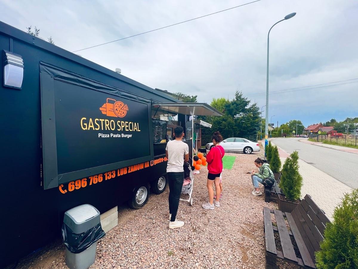 Czarny food truck z pomarańczowym logo 'Gastro Special Pizza Pasta Burger' przy drodze; klienci czekają na zamówienie, a jedna osoba siedzi na ławce.