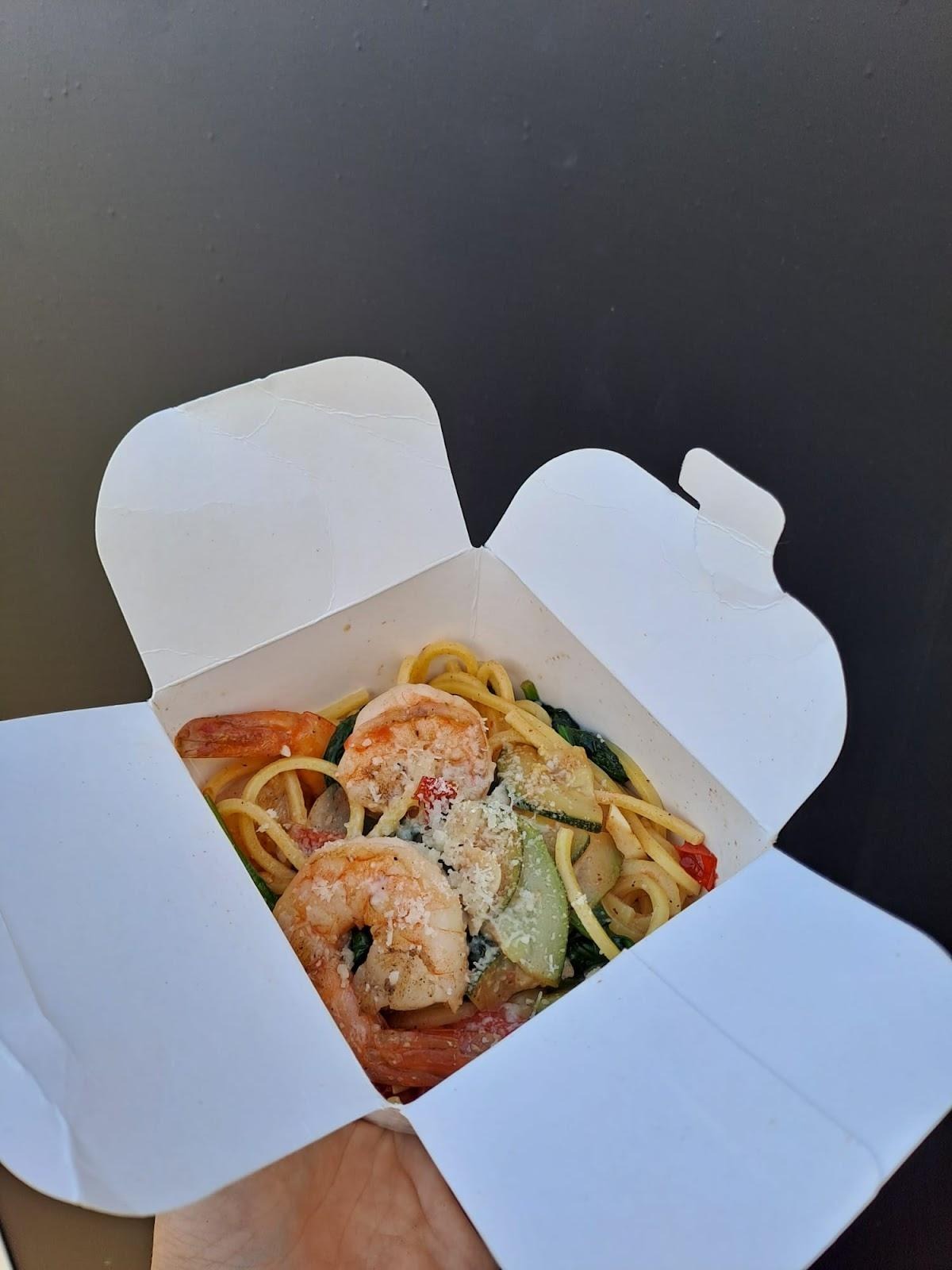 Spaghetti z krewetkami, szpinakiem i cukinią posypane parmezanem w białym pudełku na wynos, trzymane w ręce na tle czerni.