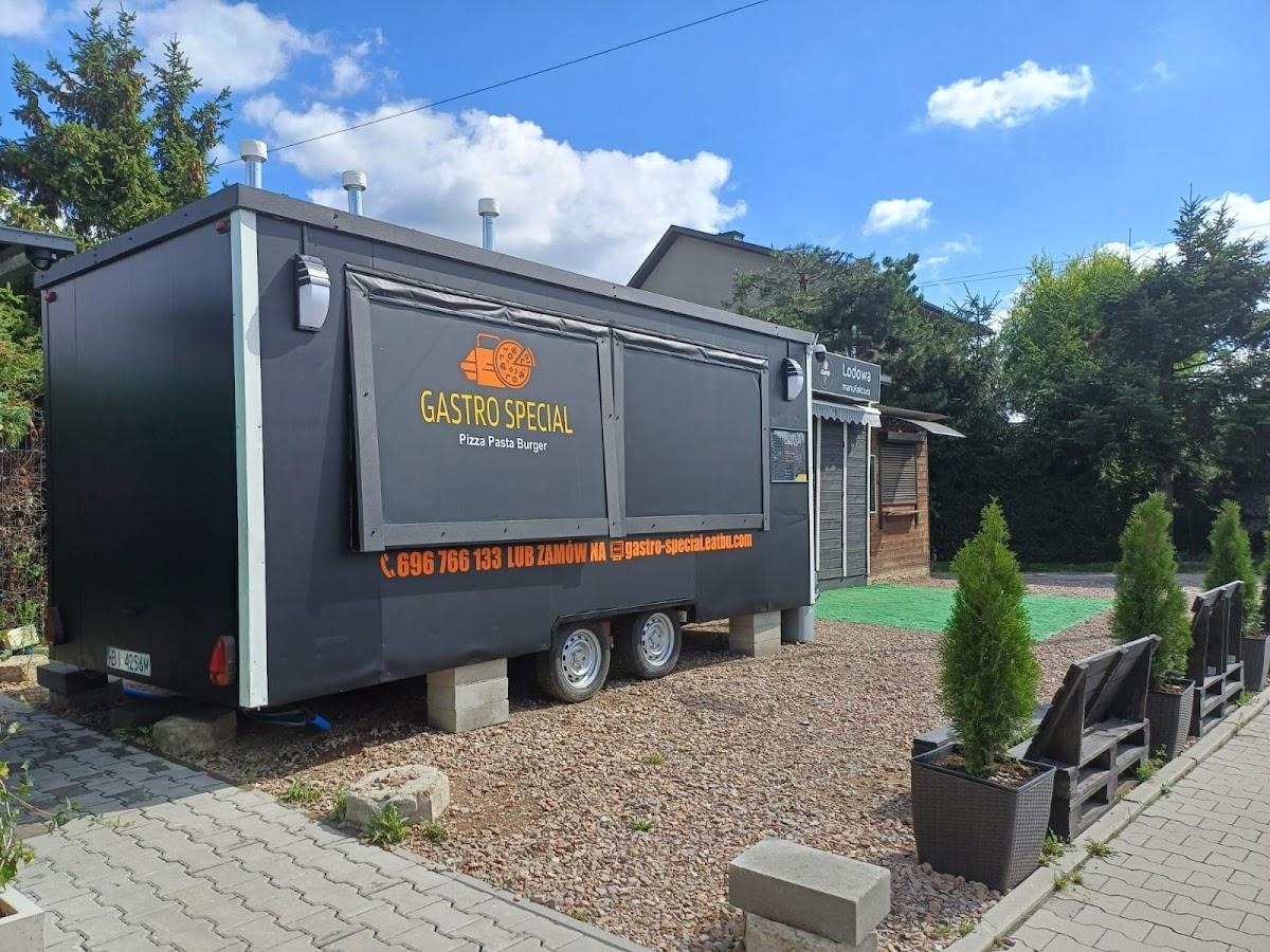 Czarny food truck 'Gastro Special' z ofertą pizza, pasta i burger, ustawiony na kostkach betonowych na żwirowym podłożu, obok budka z lodami i małe drzewka w donicach.